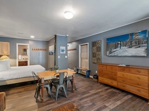 Una habitación de hotel moderna con una cama, una mesa de comedor con sillas, un aparador, un cuadro y un baño con espejo y lavamanos.