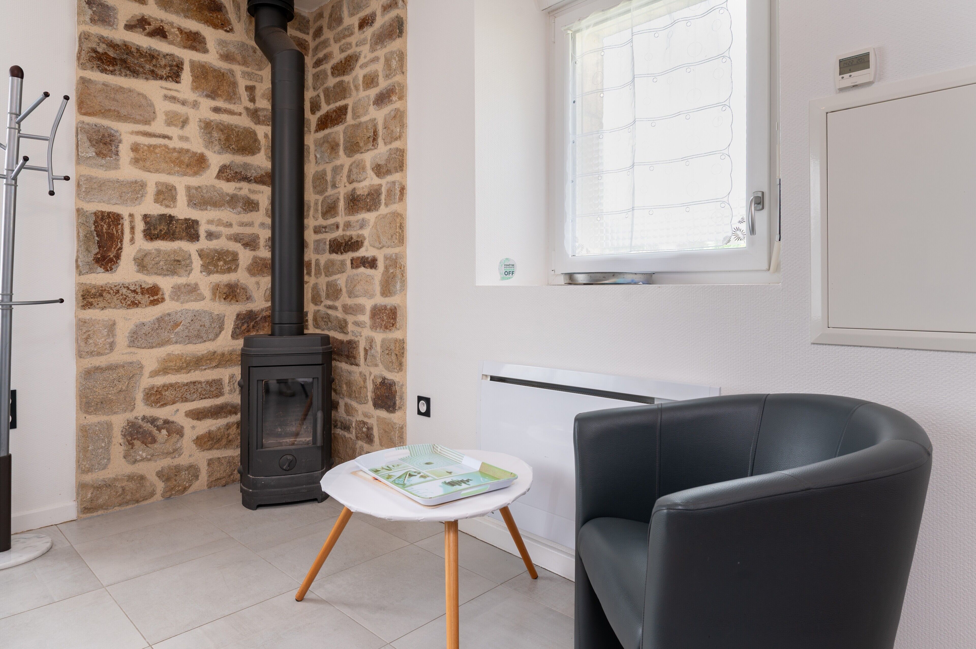 A room with a stone wall, a black wood-burning stove, a white radiator, a black leather chair, and a small white table with a magazine.