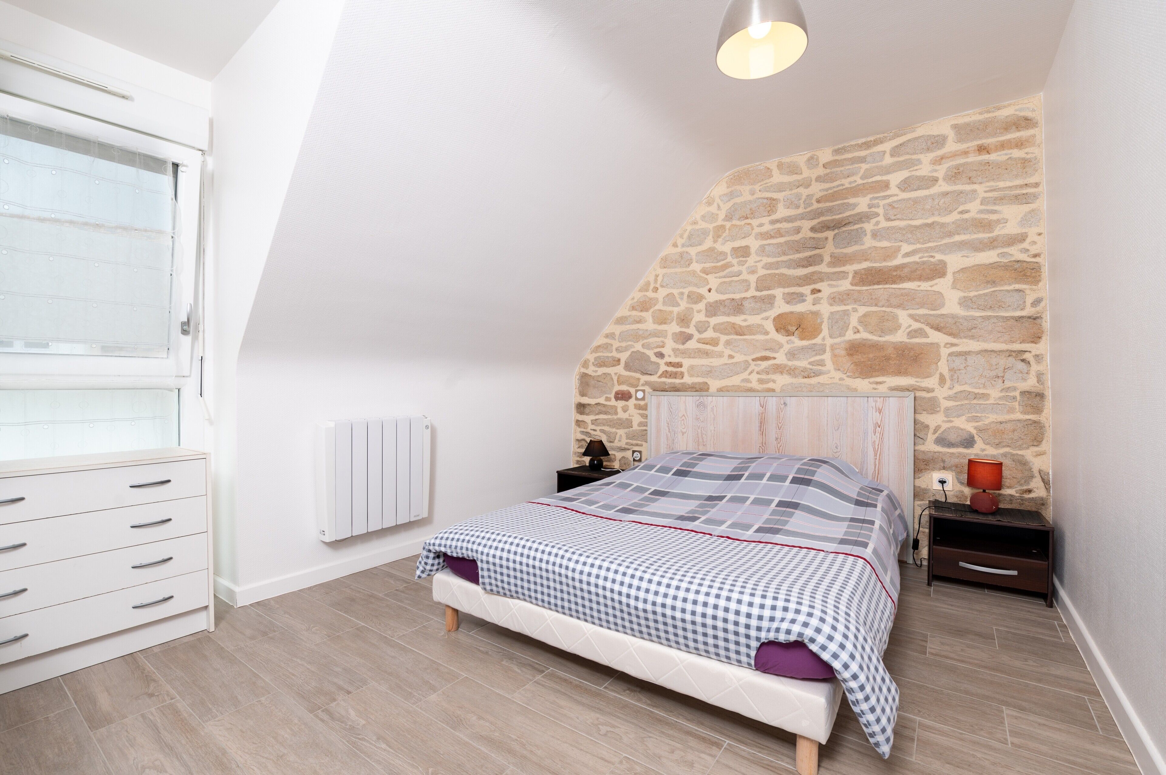A bedroom with a stone wall, a bed with a checkered bedspread, a nightstand with a lamp, and a window with blinds.