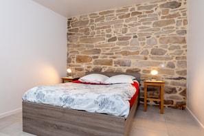 Un dormitorio con pared de piedra, cama de madera, dos mesitas de noche con lámparas y una colcha roja.