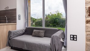 A modern room with a grey sofa, a wooden accent wall, a window with curtains, and a kitchen area. - Holiday Home 'La Longère Du Cosquer' with Private Terrace, Garden & Wi-Fi (Baden)