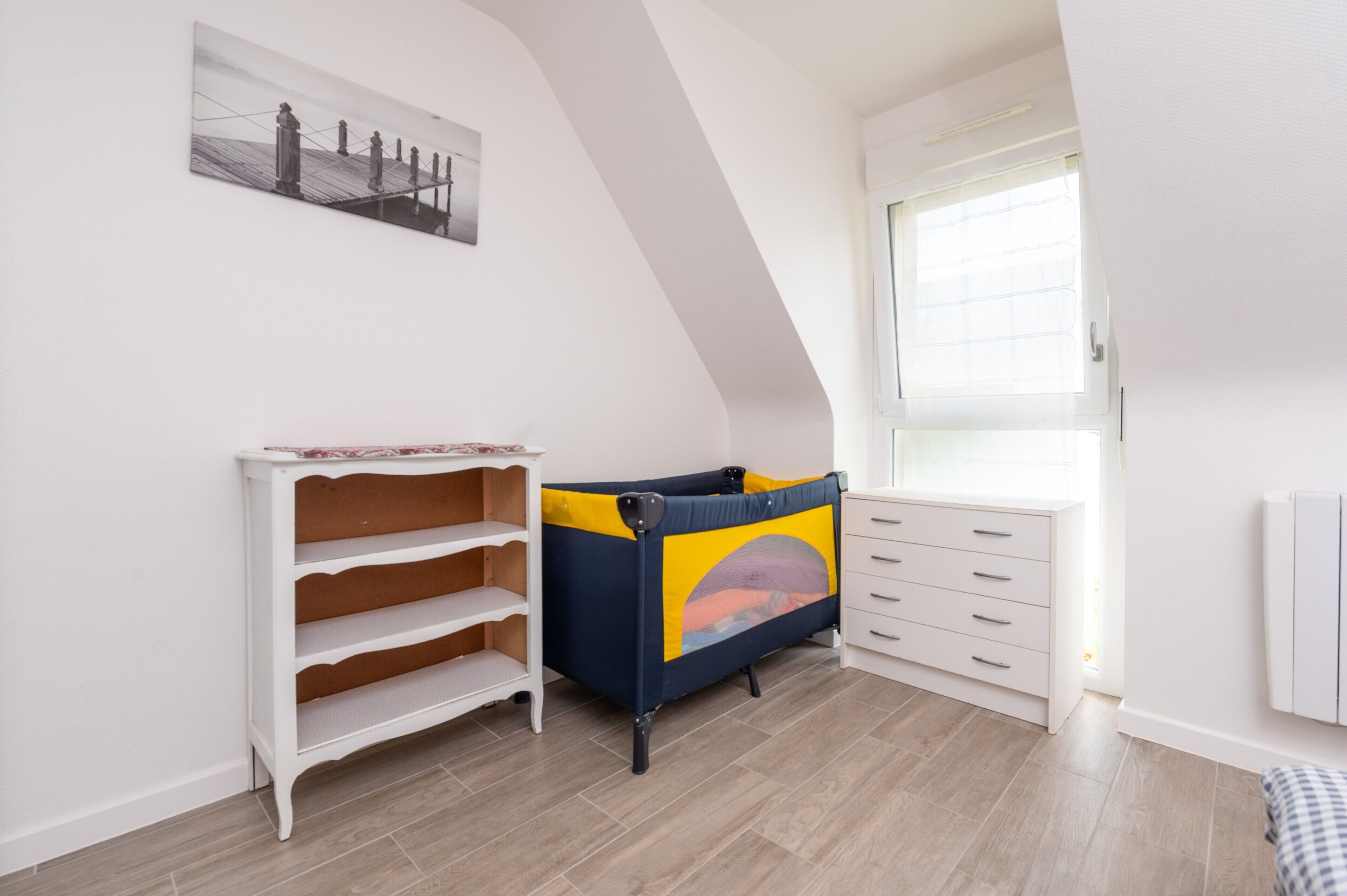 Ein Zimmer mit schrägen Wänden, einem Fenster mit Jalousien, einem weißen Schminktisch, einem mehrstufigen weißen Regal und einem bunten Spielbereich.