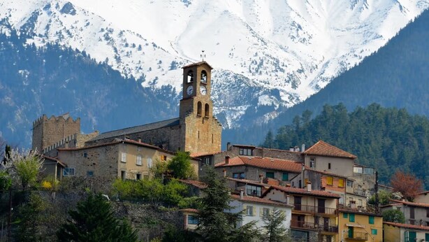 Un pueblo histórico con una imponente torre de reloj, arquitectura tradicional y montañas nevadas al fondo.