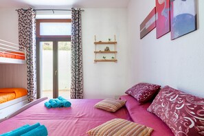 Una habitación con literas, una cama con colcha rosa, una ventana con cortinas y un estante en la pared con objetos decorativos.