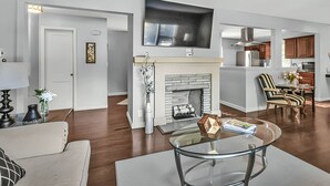 A modern living room with a fireplace, a flat-screen TV, and a dining area with a kitchen in the background. - Stay in a charming 2-bedroom house in Omaha with WiFi, AC, and a peaceful vibe. (Omaha)