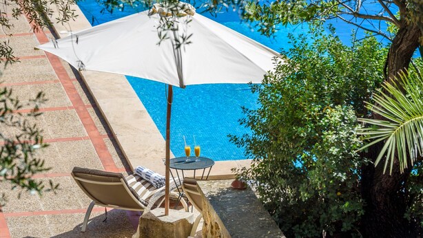 Un espace aménagé au bord de la piscine, avec un parasol blanc, des chaises longues et une table où sont servis des boissons.