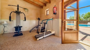 Une pièce équipée d’un tapis de course, d’un vélo d’appartement, d’une machine de musculation, d’une petite table avec une carafe, et d’une fenêtre donnant sur une piscine.