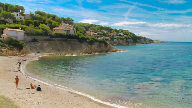 A beach with houses on a cliff, clear water, and a person walking.