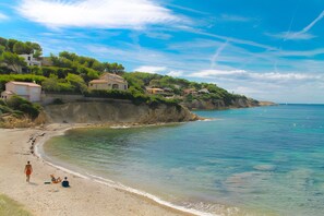 A beach with houses on a cliff, clear water, and a person walking.