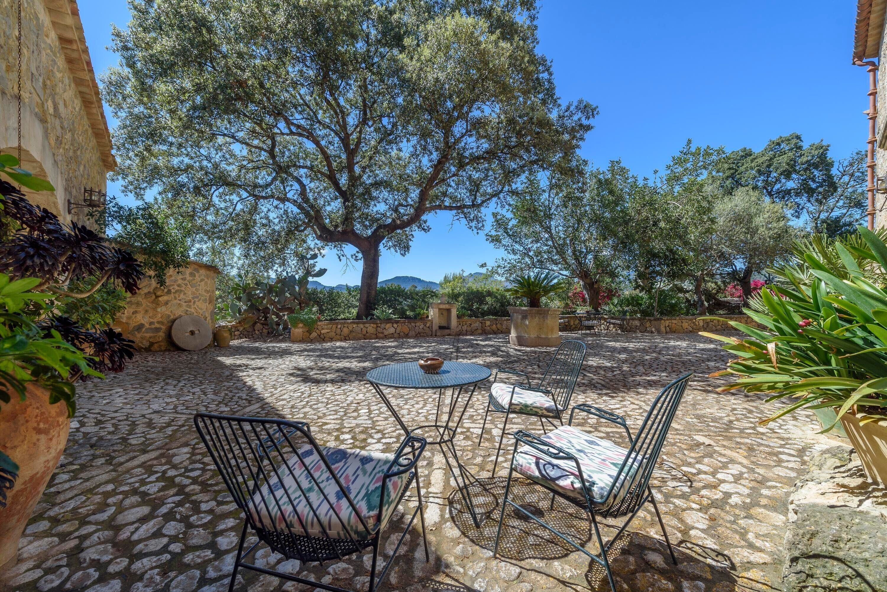 A patio with stone flooring, metal chairs, a round table, and a large tree providing shade.