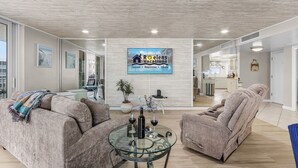 A spacious living area with a modern design, featuring a large glass coffee table, comfortable seating, and a visible kitchen area in the background.