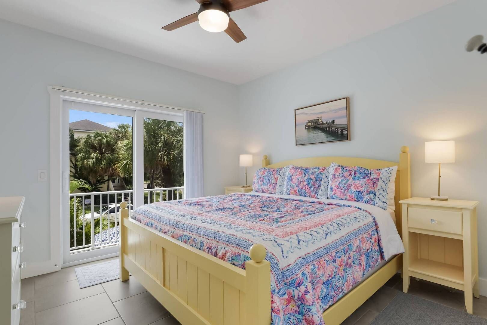 A bedroom with a bed, a nightstand, a ceiling fan, a window with a view of palm trees, and a painting on the wall.