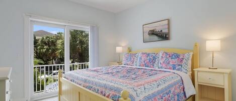 A bedroom with a bed, a nightstand, a ceiling fan, a window with a view of palm trees, and a painting on the wall.