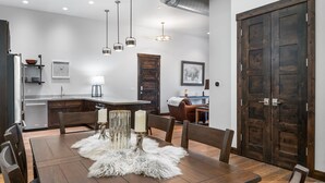 A dining area with a wooden table, chairs, and a white furry rug. There are hanging pendant lights and a wooden door in the background. - New! Downtown Whitefish with private hot tub and balcony (Whitefish)