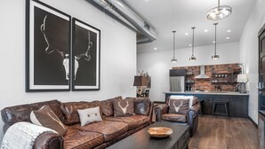 A modern living room with a leather sofa, a wooden coffee table, and a kitchen area with a brick backsplash and stainless steel appliances. - New! Downtown Whitefish with private hot tub and balcony (Whitefish)