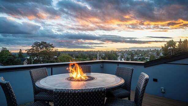 Une table ronde avec un foyer au centre, entourée de chaises en osier, sur une terrasse offrant une vue sur le paysage urbain et un ciel au coucher du soleil.