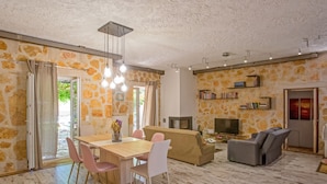 A spacious living area with a wooden dining table, pink chairs, a beige sofa, and a gray recliner. There is a bookshelf, a fireplace, and a TV on a stand. - Your Luxury Escape in Corfu’s Tranquil Countryside (Akrokefalo Gouvia)