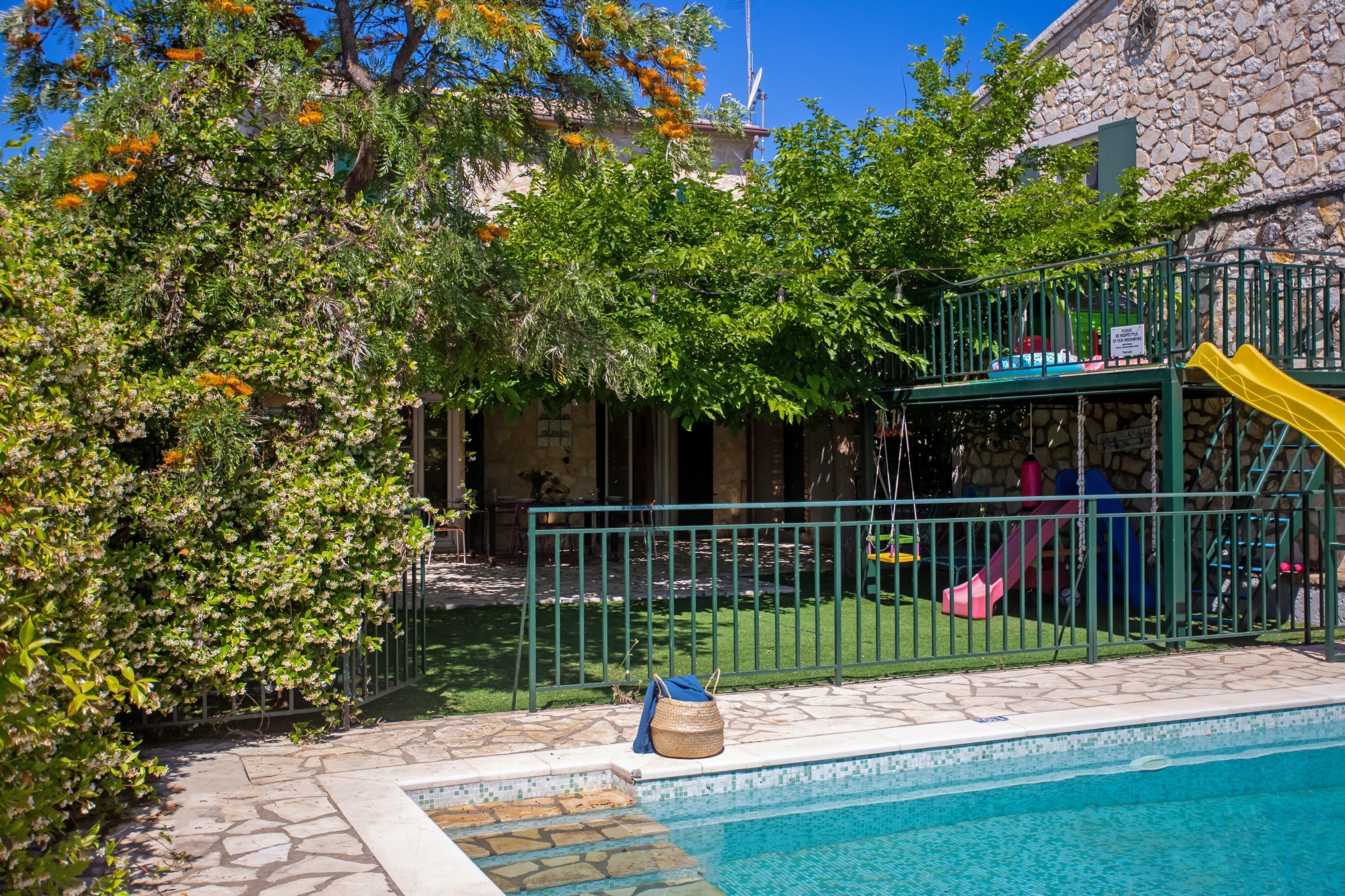 A swimming pool with a stone border, a fenced play area with a slide, and lush greenery.