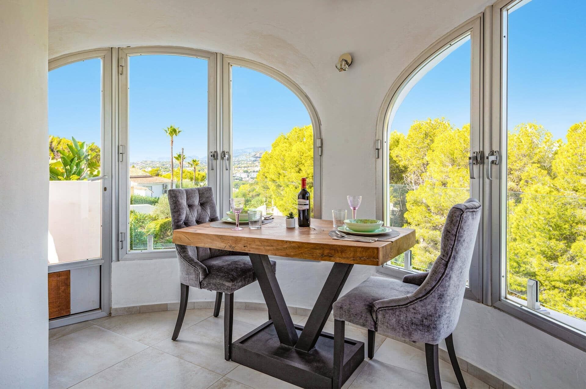 Un coin repas avec une table en bois et deux chaises grises à revêtement rembourré, de grandes fenêtres en arc de cercle et une vue sur la verdure et les bâtiments.