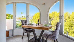 A dining area with a wooden table and two grey upholstered chairs, large arched windows, and a view of greenery and buildings. - Villa Kmar by Villa Plus (El Poble Nou de Benitatxell)