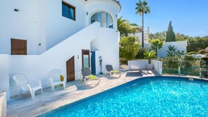 A poolside area with a clear blue pool, white chairs, and a white building with a curved balcony. - Villa Kmar by Villa Plus (El Poble Nou de Benitatxell)