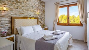 A bedroom with a brick-patterned wall, a wooden headboard, a bed with white linens, a nightstand with a lamp, and a window with curtains. - Villa Kmar by Villa Plus (El Poble Nou de Benitatxell)