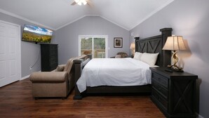 A bedroom with a bed, a chair, a nightstand, a lamp, a ceiling fan, a window, and a painting on the wall. - Hot Tub Soaks Under the Stars, Firepit S'mores & Secluded Mountain Vibes - A Country Retreat (Gatlinburg)