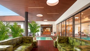 A modern indoor pool area with a wooden deck, green cushioned seating, and a view of the pool through large windows.