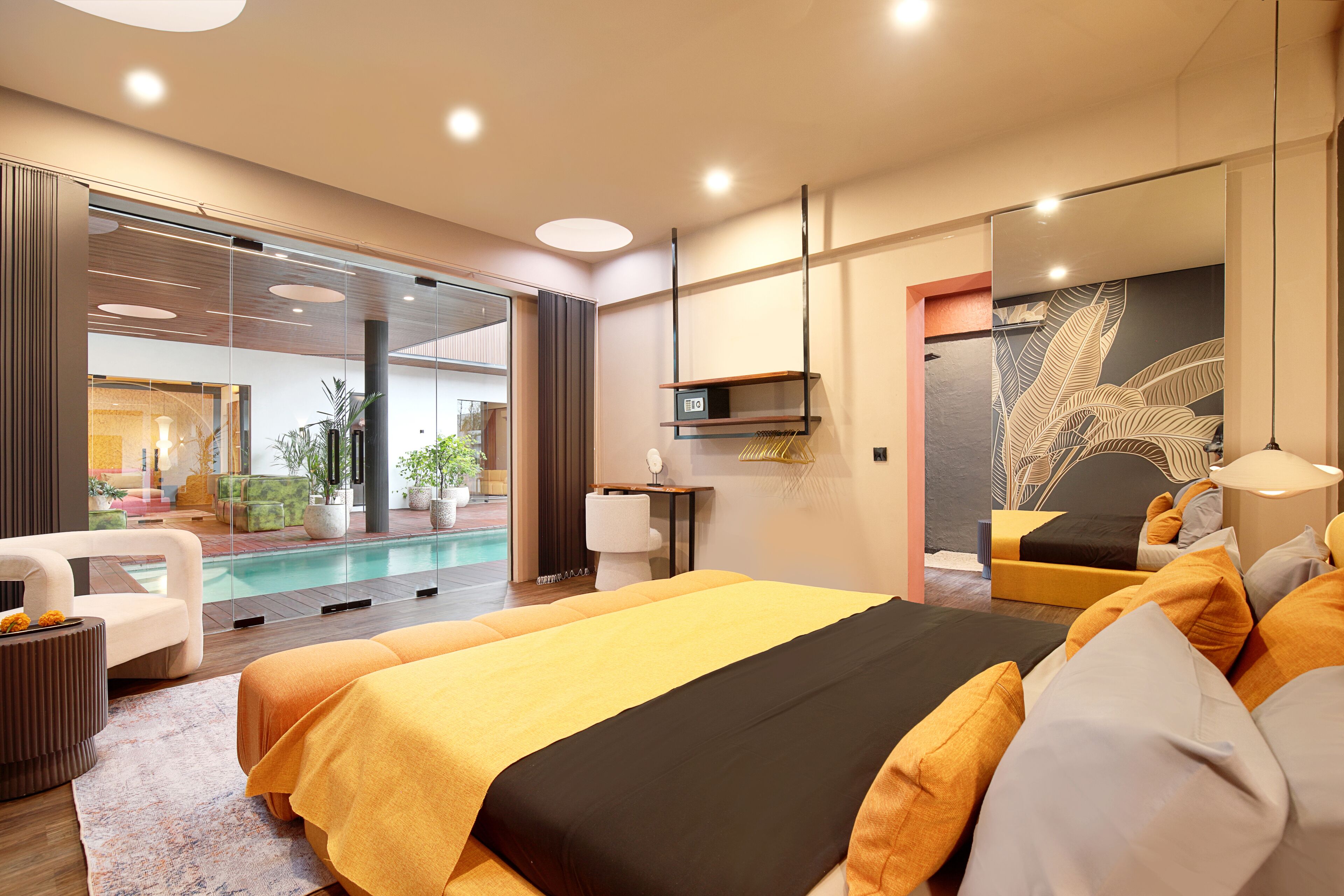 A modern hotel room with a large bed, a small seating area, a pool view, and a bathroom with a large mirror and artwork.