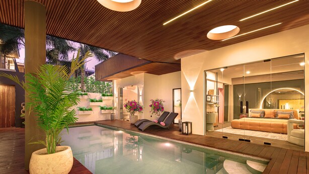 A modern indoor pool area with a wooden deck, lounge chairs, and a glass-enclosed living area with a sofa and decorative plants.