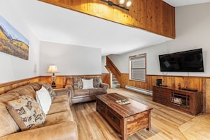 A living room with a wooden ceiling, a sofa set, a coffee table, a TV, and a staircase.