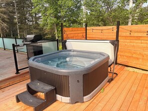 Ein Whirlpool auf einer Holzterrasse mit Glasumrandung, ein Grill und ein Holzzaun im Hintergrund.