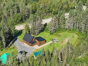 Ein zweistöckiges Haus mit geneigtem Dach, umgeben von dichtem Wald, einer sichtbaren Auffahrt und einer kleinen Außenbereich-Sitzgelegenheit.