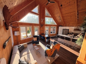 A spacious cabin with a high ceiling, wooden walls, a dining area, a kitchen, and large windows.