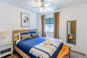 Ein Schlafzimmer mit Bett, Deckenventilator, Fenster mit Vorhängen, Nachttisch mit Lampe und einem gerahmten Spiegel.