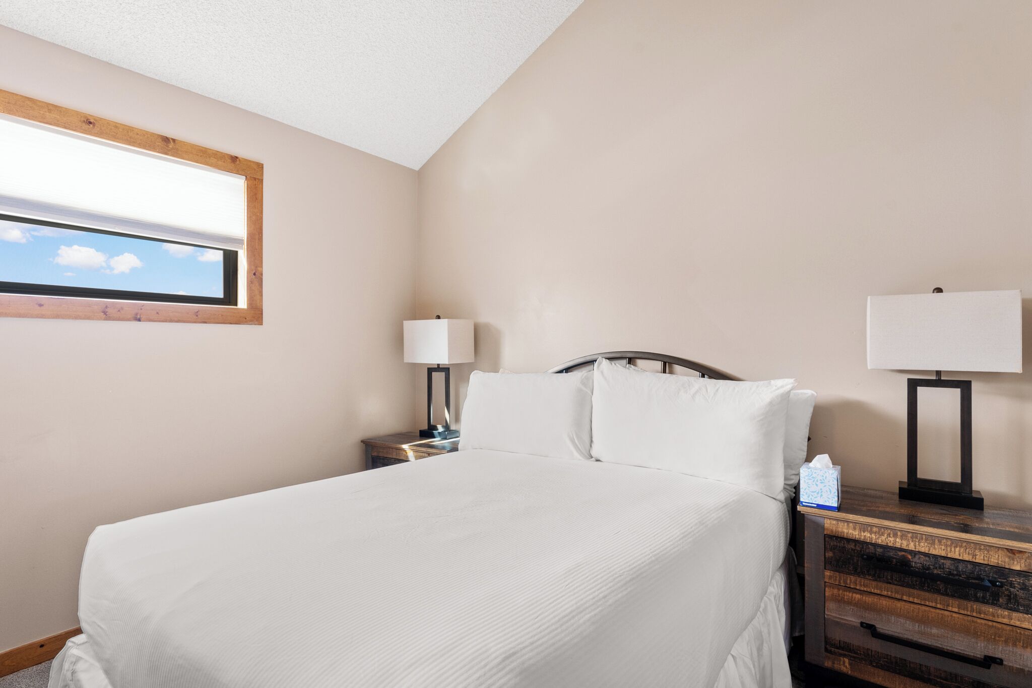 A neatly made bed with white linens, a wooden headboard, and a nightstand with a lamp and tissues.