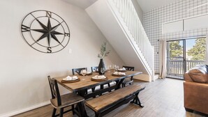 A dining area with a wooden table set for six, a black compass wall decoration, and a staircase with a white railing. - Lux Condo on Lake Conroe, Lakeview, Pool, Game room (Montgomery)