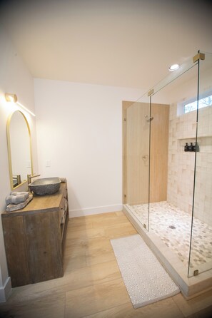 Een moderne badkamer met een houten wastafelmeubel, een grote spiegel, een douche met glazen wanden en een vloerkleed met een patroon.