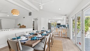 A modern dining area with a long wooden table set for six, white chairs, and a ceiling fan. - Shore Thing | Luxe Pool Oasis, Hot Tub, Siesta Key Beach (Siesta Key)