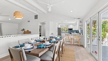 Un espace de salle à manger moderne, avec une longue table en bois pouvant accueillir six personnes, des chaises blanches et un ventilateur de plafond.