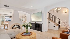 A spacious living room with a fireplace, a large flat-screen TV, and a staircase with a black railing. - Luxe Two Story Condo in the Heart of Downtown SB! (Santa Barbara)