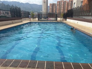 Ein Swimmingpool mit klarem, blauem Wasser, umgeben von einem schwarzen Zaun, mit Blick auf hohe Gebäude im Hintergrund.