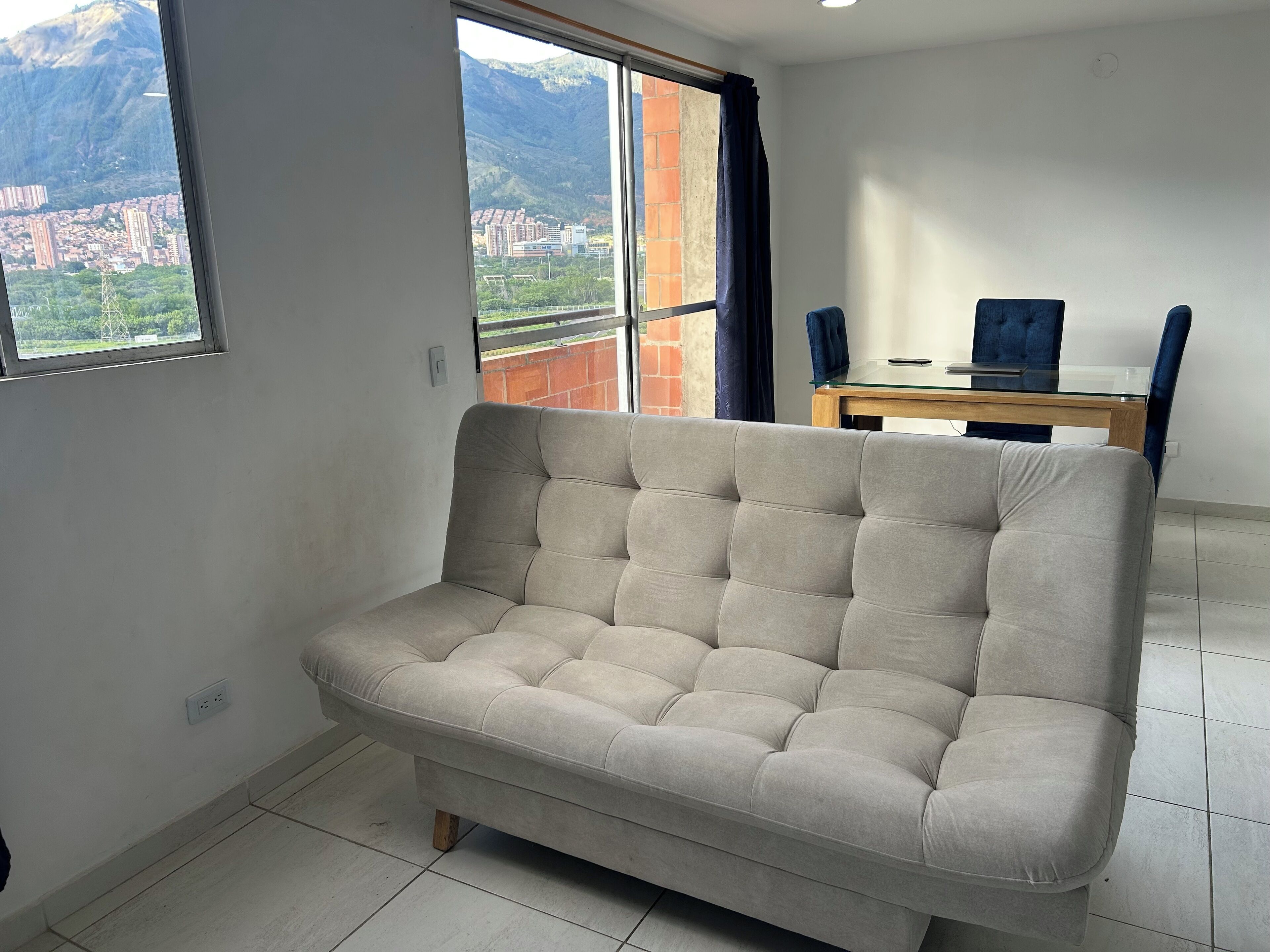 A room with a beige sofa, a dining table with chairs, a window with a view of buildings and mountains, and a tiled floor.