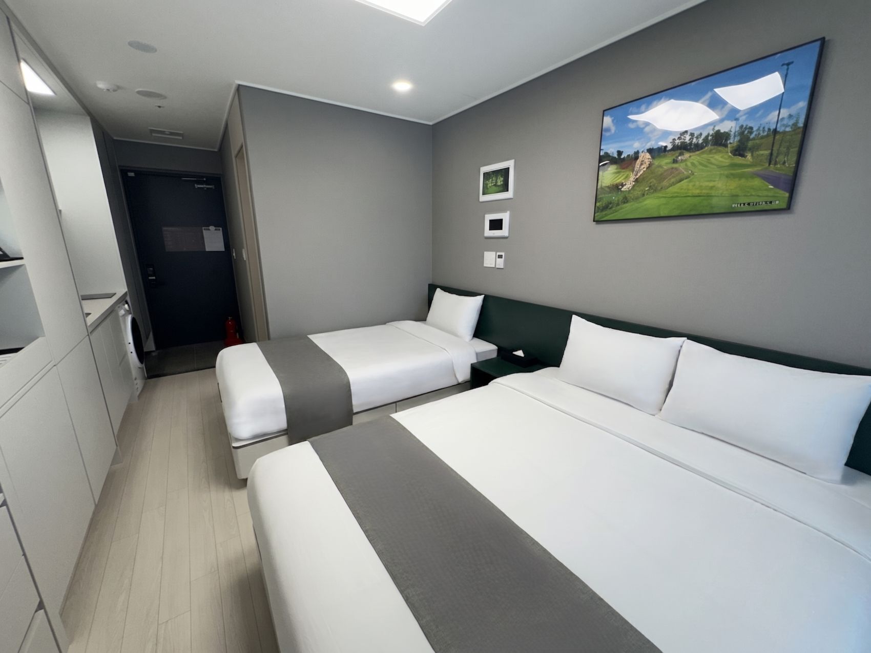 A hotel room with two beds, a washing machine, a small desk, and a picture of a golf course on the wall.