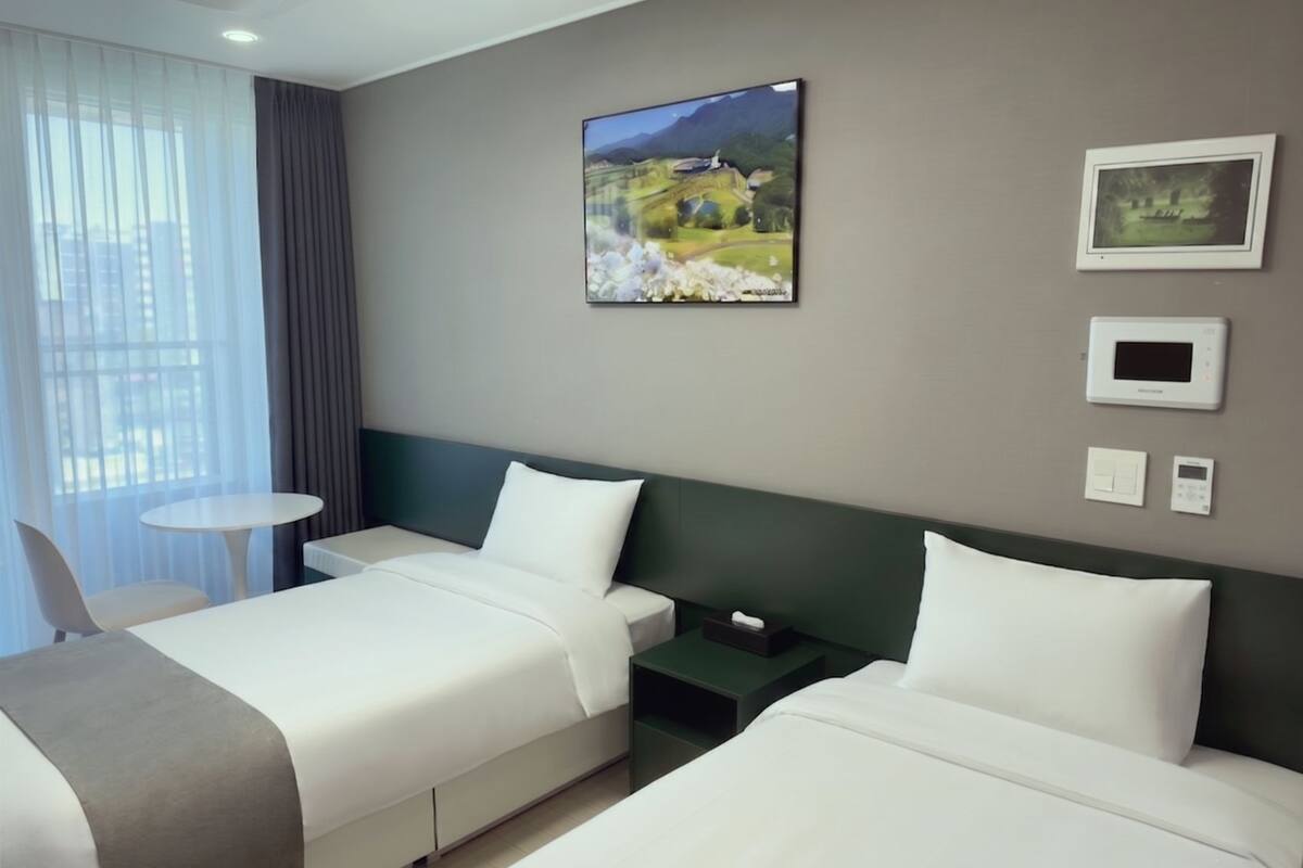 A hotel room with two beds, a small table, a chair, a painting, and a thermostat.