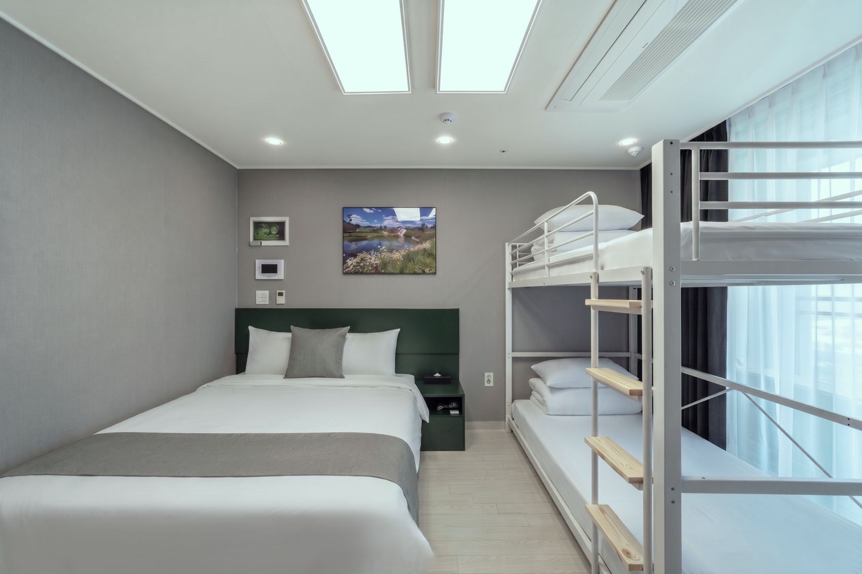 A hotel room with a bunk bed, a single bed, a nightstand, a wall-mounted TV, and a painting.