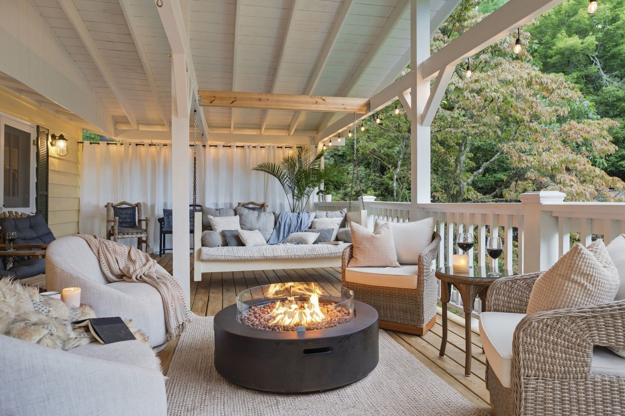 A covered outdoor seating area with a fire pit, wicker furniture, and a view of trees.