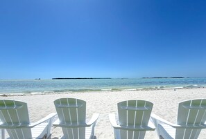 Uma praia com cadeiras brancas voltadas para o oceano, céu claro e águas calmas.