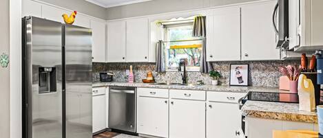 A modern kitchen with stainless steel appliances, white cabinets, a granite countertop, and a window with curtains.