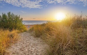 Hiekkainen polku johtaa rannalle, ja kauempana näkyy henkilö.